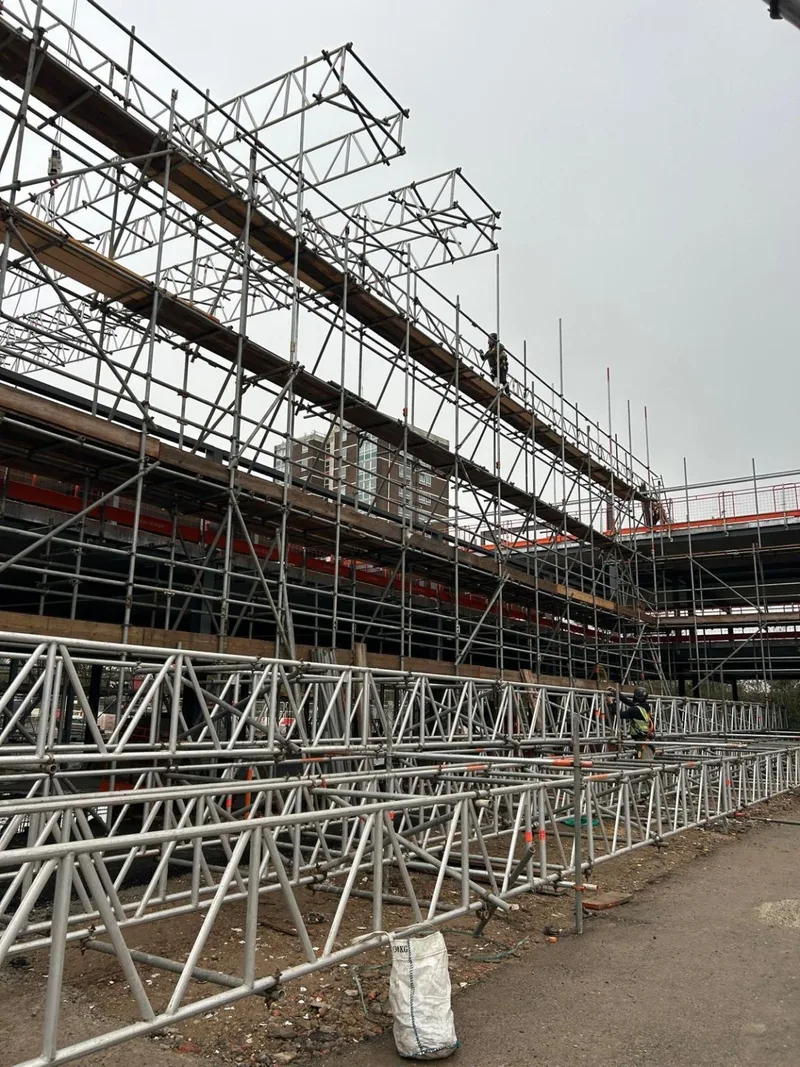 Lattice beams assembled at ground level beside the tube-and-fitting perimeter scaffold